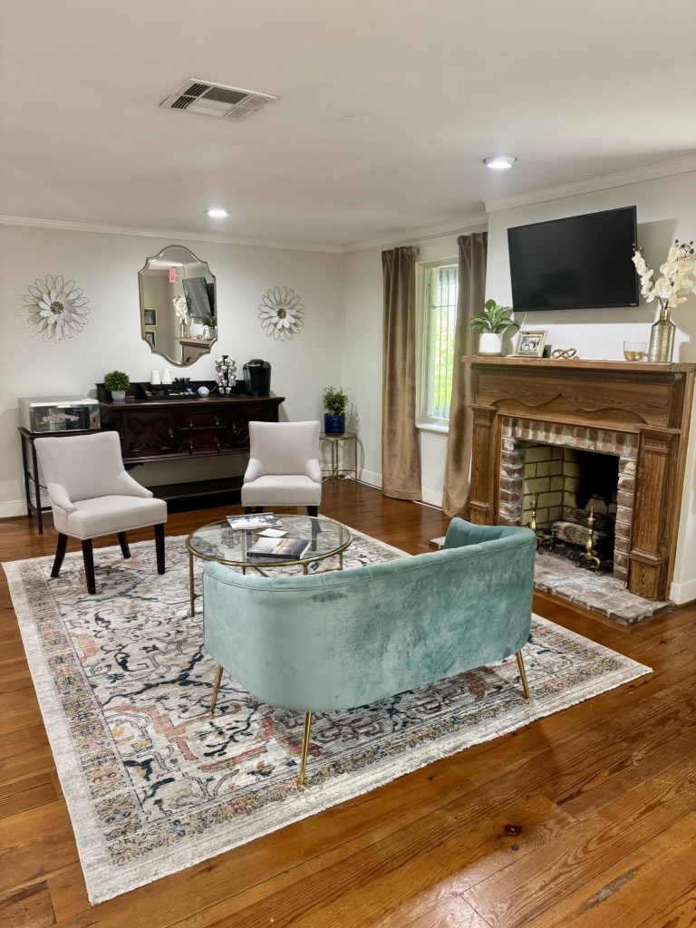 A luxurious, residential-style waiting room. It features a decorative fireplace, a light blue velvet sofa, a patterned area rug on hardwood floors, and elegant furniture, creating a relaxing, non-clinical environment