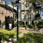 A charming exterior view of a historic multi-story building used as a dental office. In the foreground, a classic black hanging sign features a white tooth icon, surrounded by lush green landscaping and a stone pathway