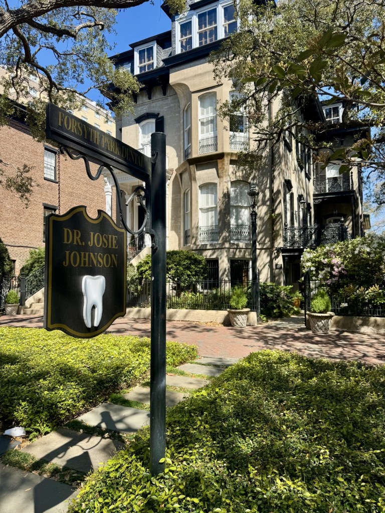 A charming exterior view of a historic multi-story building used as a dental office. In the foreground, a classic black hanging sign features a white tooth icon, surrounded by lush green landscaping and a stone pathway