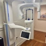 An interior shot of a modern dental treatment room featuring a high-tech 3D cone beam CT scanner (CBCT). The room has warm hardwood floors, blending advanced technology with a comfortable atmosphere