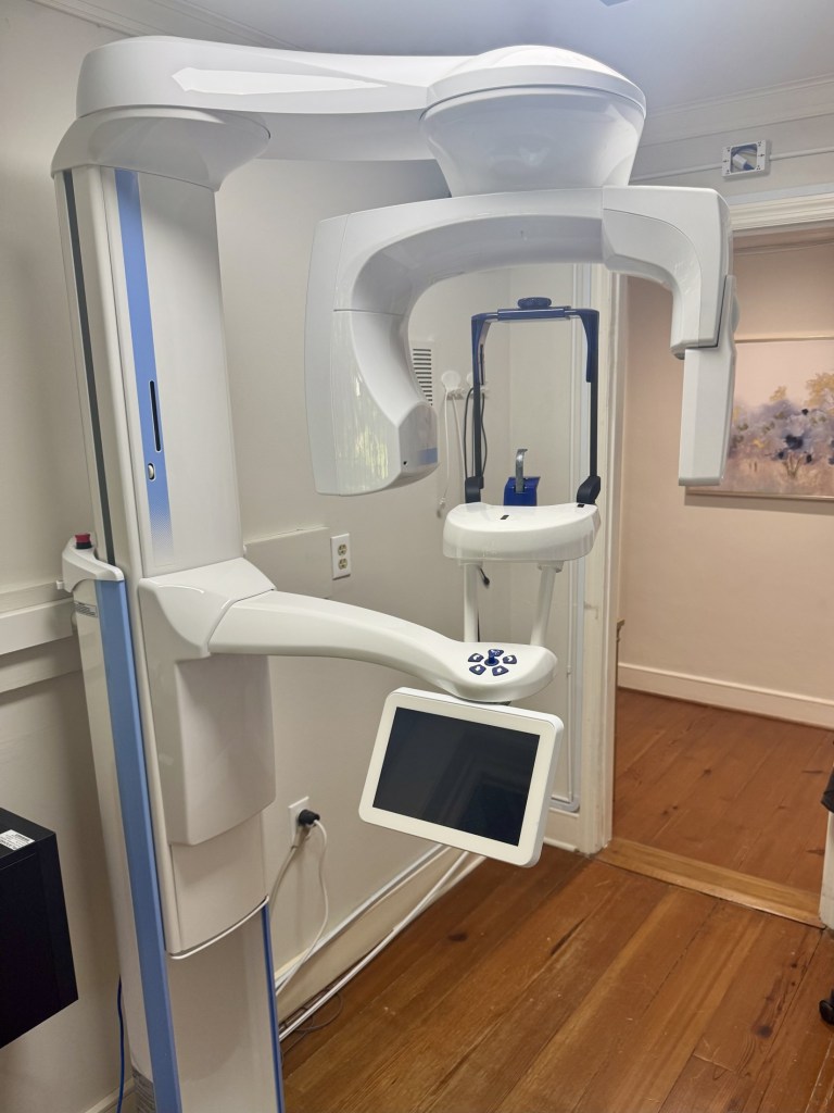 An interior shot of a modern dental treatment room featuring a high-tech 3D cone beam CT scanner (CBCT). The room has warm hardwood floors, blending advanced technology with a comfortable atmosphere