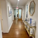 A bright, clean interior hallway with hardwood floors. A gold-framed hexagonal mirror hangs above a console table displaying professional dental products, leading toward a glass-paned office door