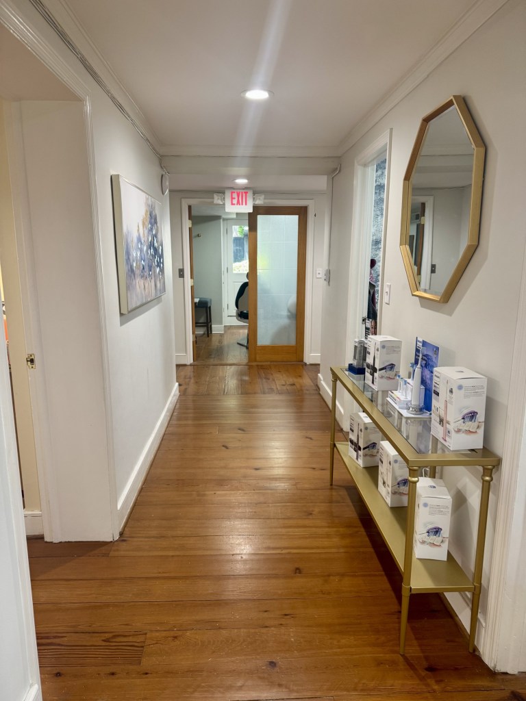 A bright, clean interior hallway with hardwood floors. A gold-framed hexagonal mirror hangs above a console table displaying professional dental products, leading toward a glass-paned office door